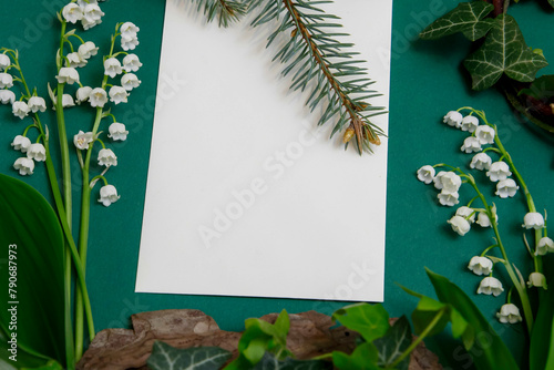 Composition on a table with white blank sheet of paper, surrounded by ivy plants and lilies of the valley. © Katarzyna