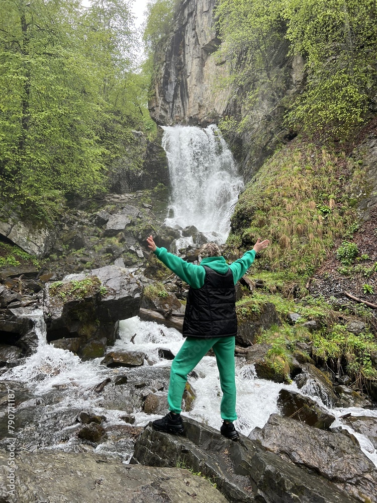 A woman on the background of the Lyazhginsky waterfall in the spring forest. A cascading waterfall surrounded by greenery. Splashes of water. The water flows along the Lyazhgi River. Ingushetia