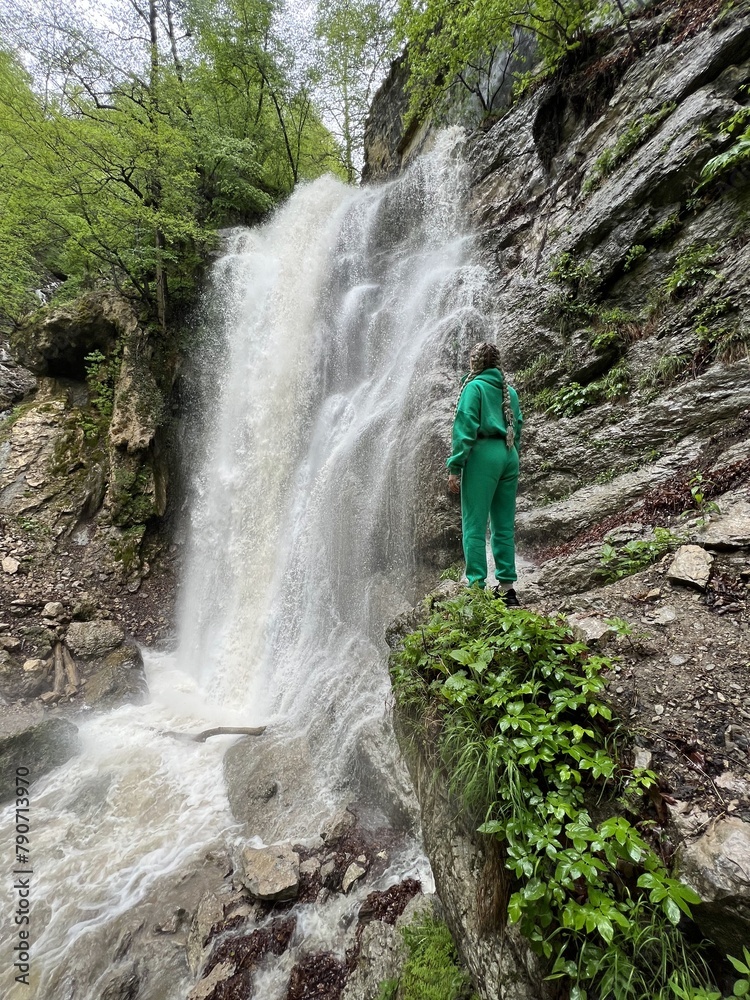 Obraz premium A woman on the background of the Lyazhginsky waterfall in the spring forest. A cascading waterfall surrounded by greenery. Splashes of water. The water flows along the Lyazhgi River. Ingushetia