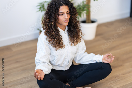 Sporty young woman doing yoga exercises while staying in lotus position in living room at home.