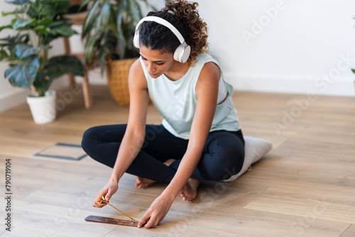Sporty young woman doing yoga exercises while listening to music with headphones and lighting incense in living room at home.