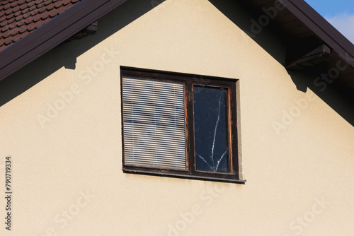 Attic window with dilapidated cracked wooden frame and closed window blinds on left and broken glass patched with transparent tape on right side