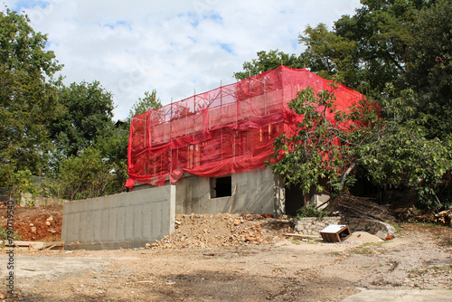 Urban family house under construction with metal building scaffolding on top wrapped with red protective mesh surrounded with gravel and dense trees in back