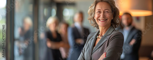 Confident senior businesswoman stands with crossed arms in office, smiling, leading her team
