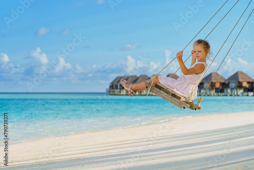  Young little girl with long blond hair swinging on a swing on the shore of the tropical sea