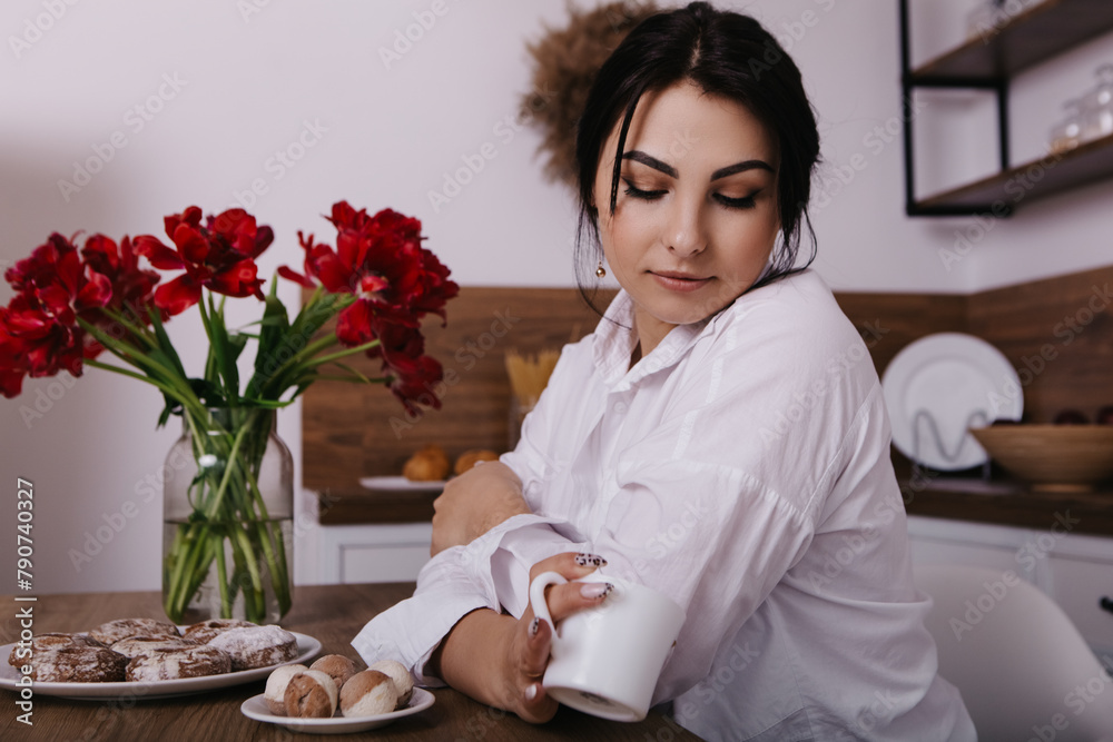 Woman poses with vibrant bouquets of red tulips in vase in kitchen, drink hot tea beverage coffee, eat sweets. Spring International Women`s Day, 8th of March. Holiday, festive, celebration Woman poses with vibrant bouquets of red tulips in vase in kitchen, drink hot tea beverage coffee, eat sweets. Spring International Women`s Day, 8th of March. Holiday, festive, celebration
