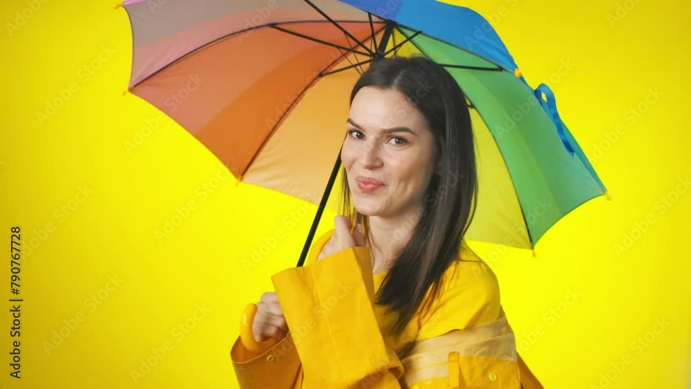 A happy attractive woman in a bright yellow raincoat with a rainbow umbrella is checking for the rain. Yellow studio background