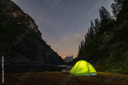 A yellow tent sits next to a large pond in the mine shaft. There is a pine forest next to the pond and a large mountain on the other side..Star shooting adventure In the dark night.water reflection