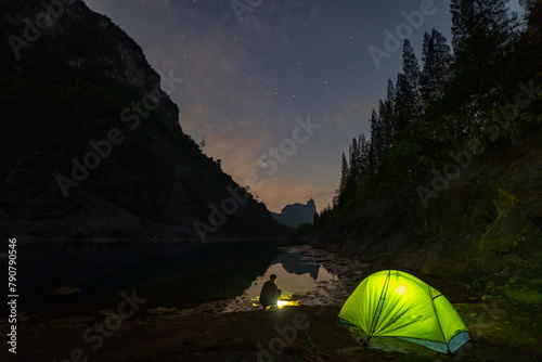 A yellow tent sits next to a large pond in the mine shaft. There is a pine forest next to the pond and a large mountain on the other side..Star shooting adventure In the dark night.water reflection
