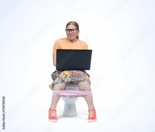 A happy young woman in glasses and a colorful skirt is sitting on the toilet with a laptop on her lap, isolated on a light blue background