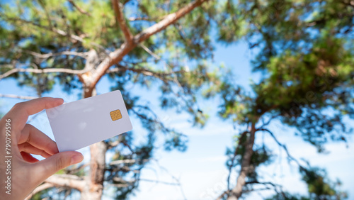 A white bank card is held in a woman's hand, set against the backdrop of a pine tree and the sky. © Jūlija