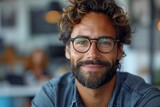 A smiling man with curly hair and glasses seated in a bright modern workplace, exuding positivity
