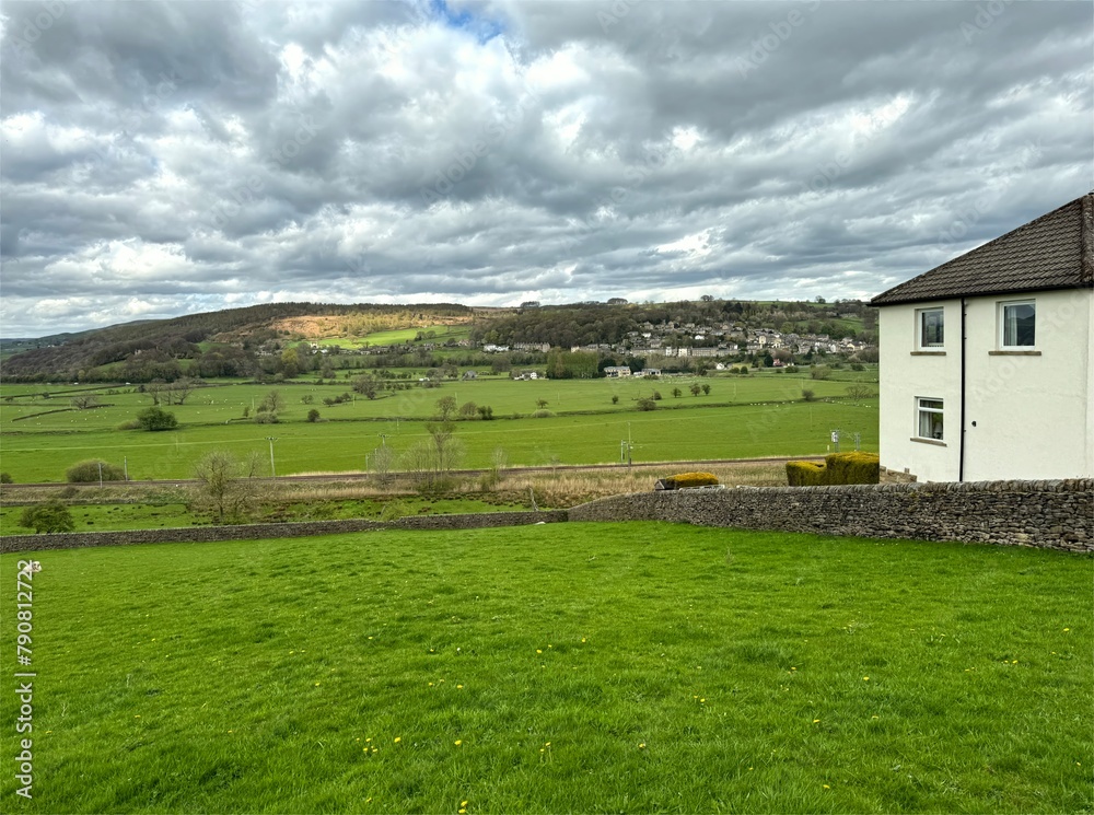 A white house sits on the edge of lush green fields in the Aire Valley. In the distance, Kildwick nestles among rolling hills, on a cloudy late winter's day near Crosshills, Yorkshire, UK.