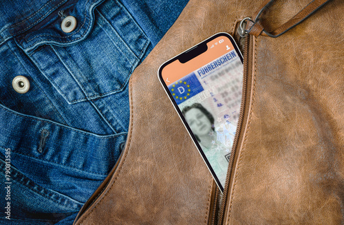 Aus einem geöffneten Reißverschluss einer braunen Handtasche schaut ein Smartphone heraus, auf dem gerade eine App mit digitalem Führerschein geöffnet ist. Im Hintergrund ist eine Jeansjacke zu sehen,