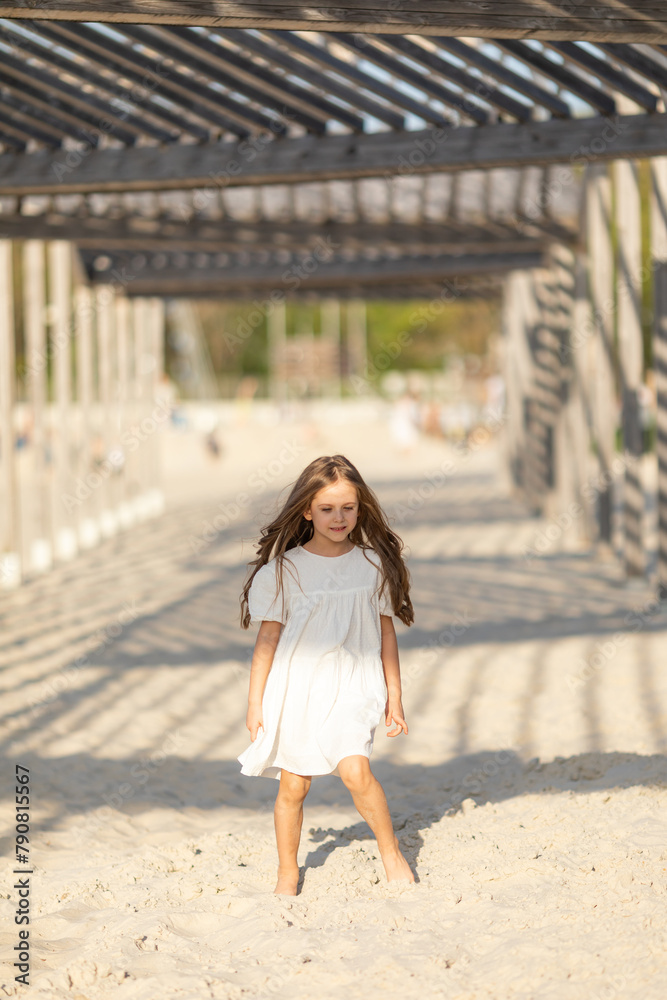 Happy little girl in white summer dress playing and has fun on a white sandy beach near the ocean in summer. Children's holidays. Earth Day. Children's Day. Vacation concept. Space for text Happy little girl in white summer dress playing and has fun on a white sandy beach near the ocean in summer. Children's holidays. Earth Day. Children's Day. Vacation concept. Space for text