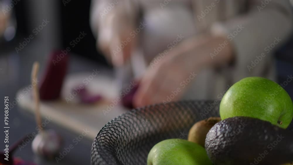 Close-up of a basket of fruits on the kitchen table. In the background, out of focus, an unrecognizable woman is cutting red beet with a kitchen knife on a wooden cutting board Close-up of a basket of fruits on the kitchen table. In the background, out of focus, an unrecognizable woman is cutting red beet with a kitchen knife on a wooden cutting board