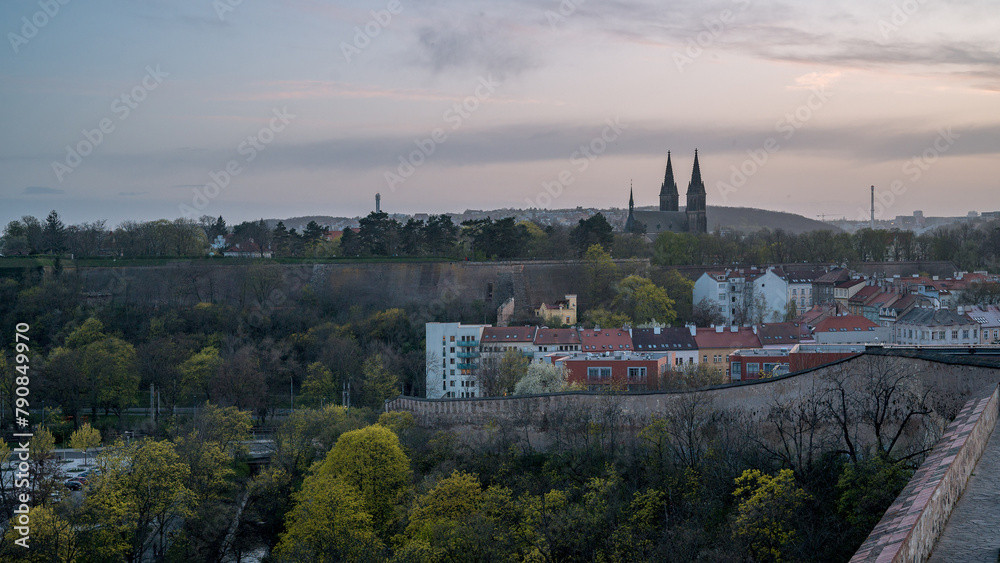 This image captures the serene sunset view from Nusle Bridge, featuring the Vyšehrad Cathedral in the distance. The air is filled with a unique glow from the Saharan dust particles, adding a mystical 