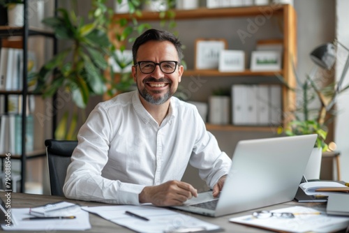Happy male employee working on laptop in modern office, sitting at desk with documents and pen smiling to camera, wearing glasses looking happy while doing work using computer for professional