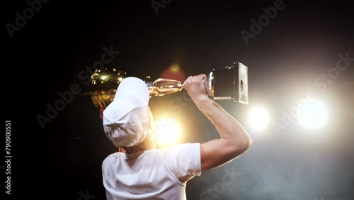 Happy winner man raising trophy with joy for sport success standing on arena stage in spotlights back view. Professional victory in competition with achieving cup