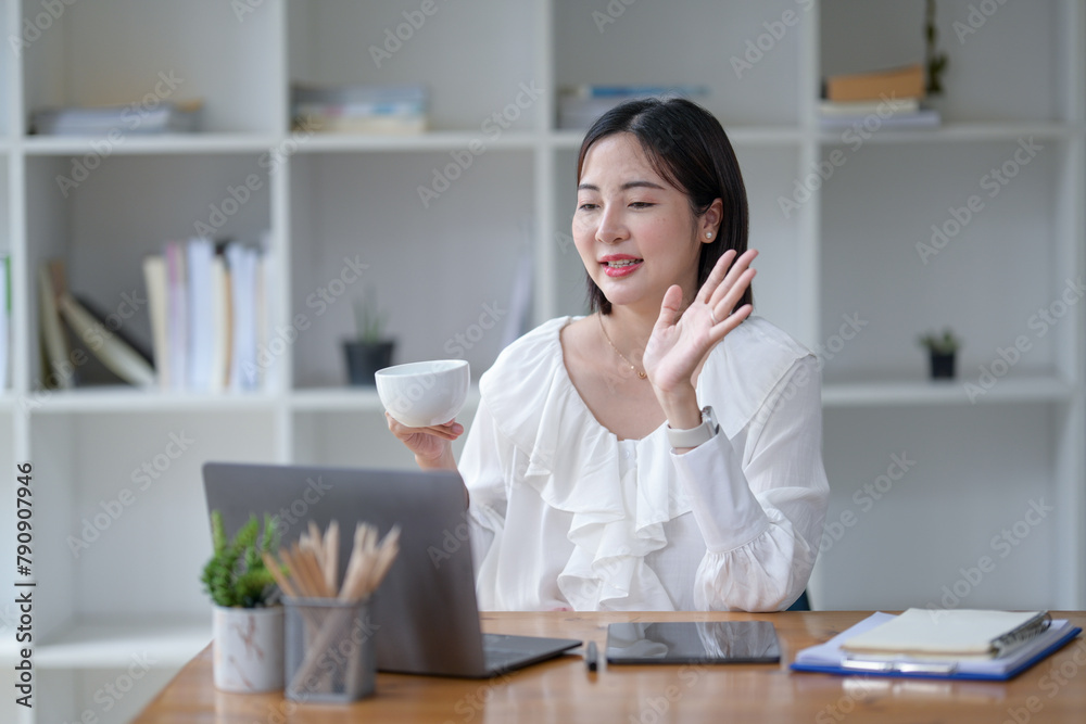 Asian woman sitting working with laptop and drinking coffee at desk in office. Talking through the laptop screen Asian woman sitting working with laptop and drinking coffee at desk in office. Talking through the laptop screen