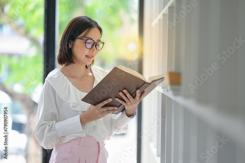 A businesswoman with glasses engrossed in reading a book at a modern office with a bookshelf.