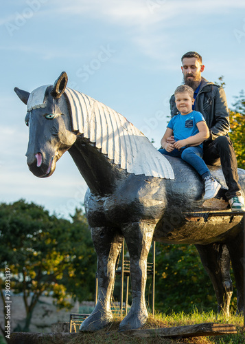 Portrait of a father and his very pleased son riding a large wooden horse in a field. Vertical photo