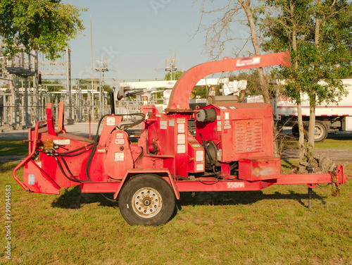 Side view of a red wood chipper in grass field of electric substation with white trucks and steel utility poles sections. Green trees and blue sky. Late afternoon sun and shade. Copy space. 
 photo