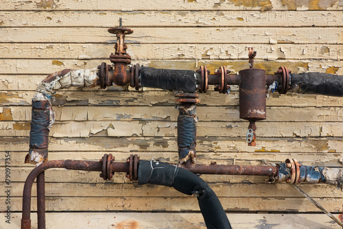 A fragment of an old rusty heating system made of pipes and valves against the background of the wall of an abandoned wooden house. Background. Grunge