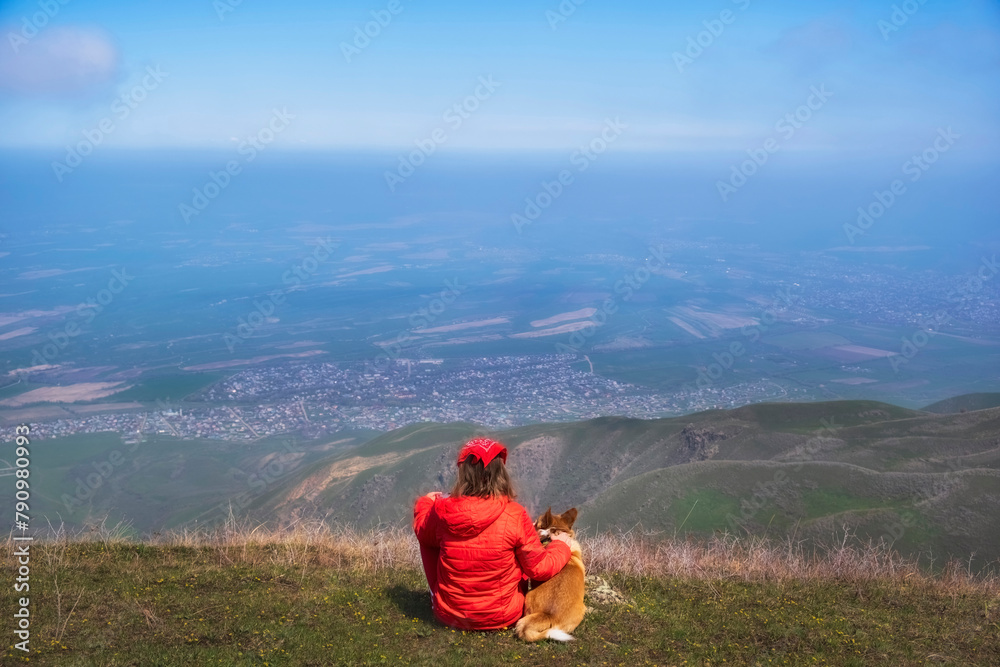 A woman and her friend, a red Corgi dog, admire the surrounding nature from a hill or mountain on a sunny spring day. A woman and her friend, a red Corgi dog, admire the surrounding nature from a hill or mountain on a sunny spring day.