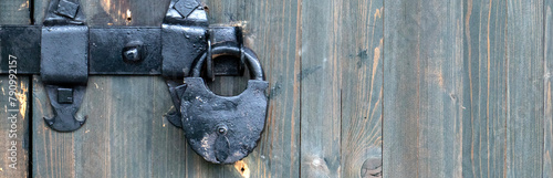 There is a rusty lock hanging on the wooden gate. Close-up view of a closed door. The wooden door is locked. A crude lock on the door, painted black. photo