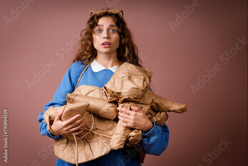 Young positive smiling woman receives surprise gift and holding in hands bunch of gifts wrapped in rustic beige craft paper. Hat, shoes, hair dryer, soft toy and glasses