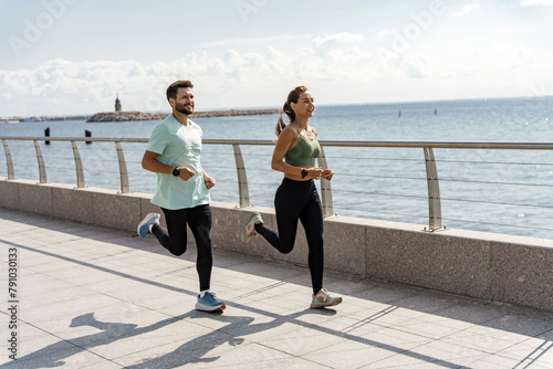 Exuberant couple enjoying a seaside jog against a backdrop of glistening waters under a clear sky. © muse studio