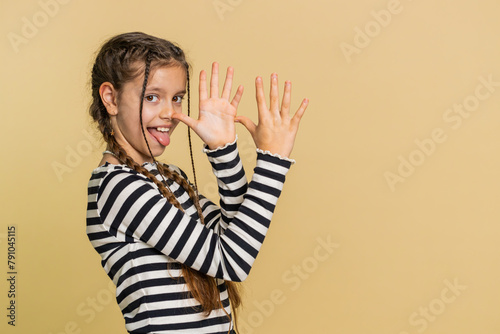 Cheerful funny young school girl showing tongue making faces at camera, fooling around, joking, aping with silly face teasing bullying, abuse. Preteen female child kid 10 years old on beige background