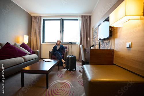 Adult caucasian businesswoman talking on smartphone in armchair in hotel room