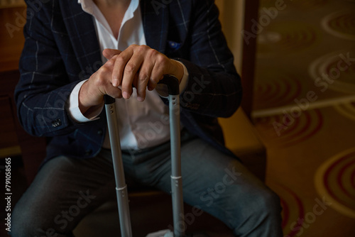 Cropped businessman with suitcase sitting on leather chair in blurred hotel room