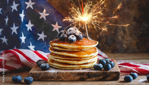 A stack of homemade American pancakes with maple syrup and blueberries on a wooden plank decorated with a sparkler and a flag of the USA in the background