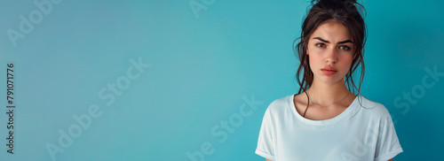 Portrait of an attractive young sad and unhappy brunette woman on a blue banner background. A sweet, upset and offended girl in a simple white T-shirt. photo
