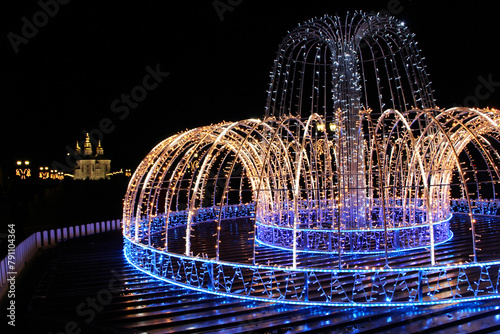 Beautiful fountain of colored holiday garlands glittering in city park in winter © alexmak