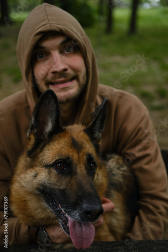 A young guy is sitting in a spring forest and hugging a German shepherd. Male owner with a dog on a walk in a green park. Front view portrait of two best friends.
