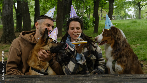 Dogs celebrates birthday with family. Young couple with Australian and German Shepherds in spring green park. Pets birthday party with meat cake and 7 candle, paper hats on heads.