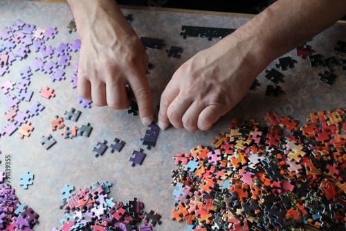 Close-up of adult caucasian hands diligently putting together a vibrant and intricate jigsaw puzzle on a wooden table surrounded by scattered puzzle pieces of various shapes and colors