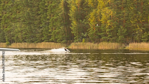 Man in wetsuit wakeboarding on serene lake, surrounded by lush green trees in peaceful forest on sunny day. Clear blue water provides stunning backdrop for thrilling adventure.