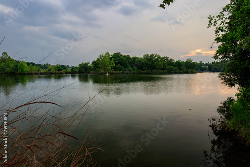 Soft ripples grace the calm lake under a cloudy sky as the sun sets. Lush trees adorn the distant shore, framing the scene in natural tranquility. In the backdrop, a solitary kayaker glides along.
