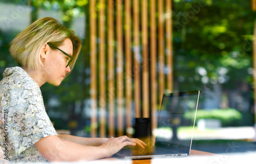 A woman in glasses outside the window is working on a laptop, a businessman is writing a message. Freelancer working away from home, sunny summer day, business project, blonde girl with short hair