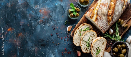 Italian ciabatta bread sliced on a wooden cutting board with herbs, garlic, and olives on a dark textured background, with space for text, seen from above.