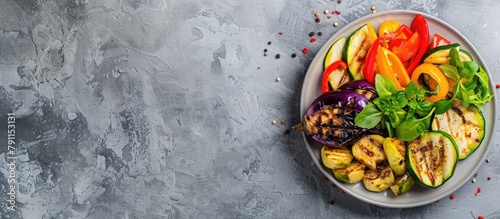 Colorful grilled vegetables including bell peppers, zucchini, and eggplant arranged on a plate placed on a light grey slate, stone, or concrete surface. Viewed from above with space for text. photo