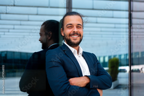 Confidence successful businessman in suit with beard standing in front of office glass building lean on wall arm crossed looking at camera and smile. Hispanic handsome modern business man portrait