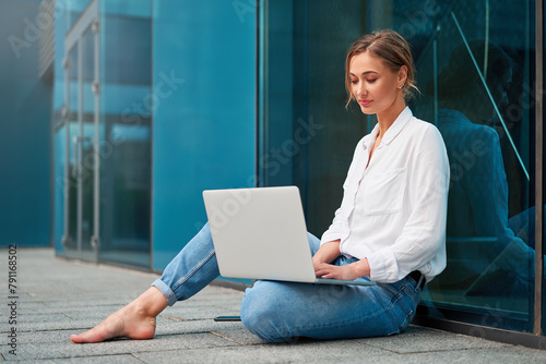 Young woman checking email on laptop sitting on the ground outside sitting barefoot outdoors on floor near modern office center