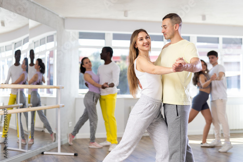 Positive young adult couple in casual sportswear enjoying slow foxtrot in sunny dance studio. Amateur social dancing concept