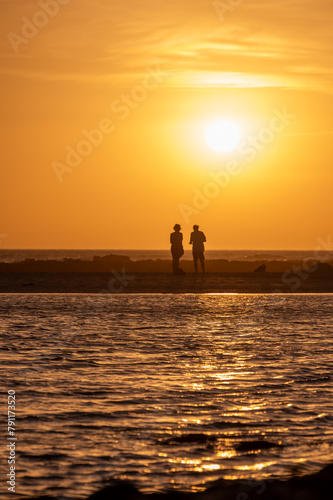 Man and woman staying and looking at sunset on ocean beach, orange sky, silhouettes of people on vacation